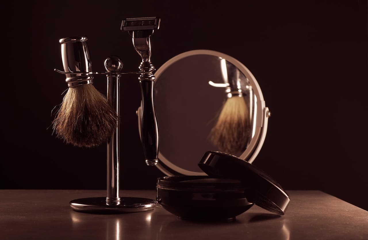Shaving brush and razor on a steel stand beside a round mirror, in low warm light